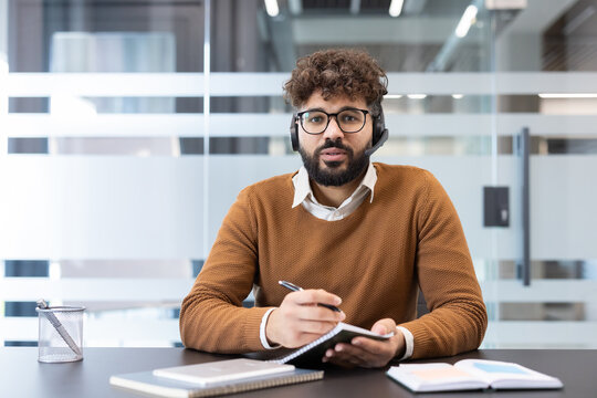 Young man wearing a headset and glasses, actively participating in a video conference from his office desk, looking at the viewer and taking notes in a notebook