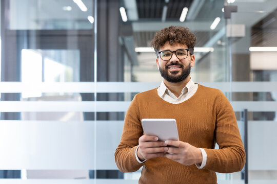 Young professional man smiling at the camera, holding a digital tablet while standing in a modern office hallway, representing technology, communication, and business success
