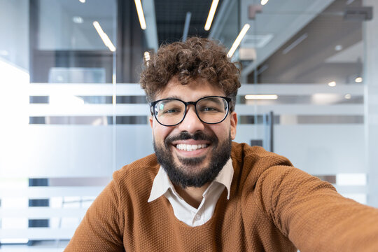 Young professional man with beard and curly hair wearing glasses, smiling brightly while making a video call or taking a selfie in a contemporary office environment