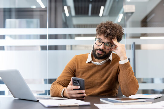 Young man with curly hair and beard sits at an office desk holding a smartphone, looking bored and confused, illustrating procrastination, distraction and workplace doubt