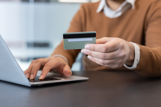 Person making an online payment, holding a credit card in one hand and typing on a laptop with the other, engaging in e-commerce and secure banking transactions from a modern office setting
