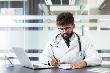 Fototapeta premium Young male doctor with curly hair and beard wearing glasses and a white lab coat with a stethoscope, sitting at a desk and writing on a clipboard in a modern clinic or hospital office