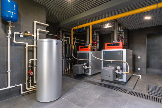 Two large boilers sit side by side in a utility room. A metal storage tank stands nearby. Pipes run along the walls and connect the equipment. The room is well-lit and features gray tiles
