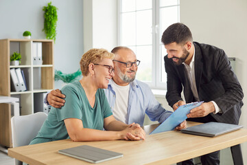 Smiling senior couple consults a financial advisor in a bright office, reviewing paperwork and discussing contract about investments and insurance going to sign documents during a meeting.  © Studio Romantic