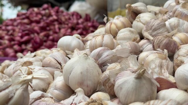 Large piles of fresh garlic bulbs are displayed for sale on a market stall, with vegetable assortments and busy trading rows visible behind. Fresh produce market.