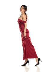Fototapeta premium Side view of a brunette woman in a red evening dress standing full body on a white background