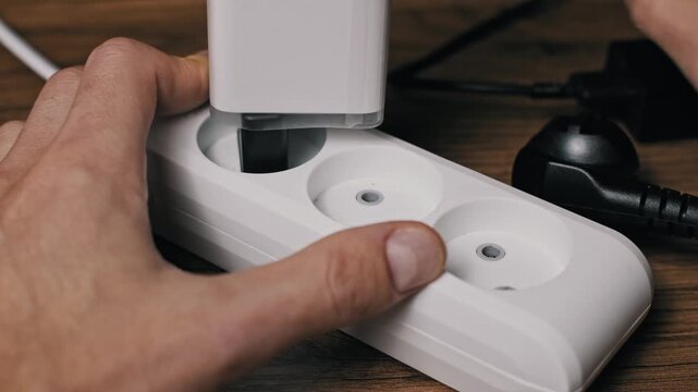 Close-up of male hands unplugging a black power cord and a white adapter from a white plastic extension strip. The sequence on a wooden table shows energy conservation and safety.