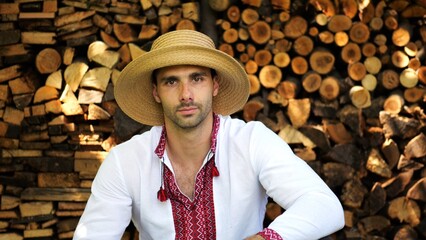 Portrait of ukrainian guy in straw hat against the background of woodshed. Young man wearing an embroidered shirt looking into camera outdoor. Concept of cultural heritage and ethnic roots