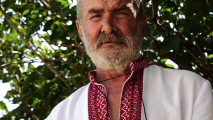 Portrait of senior ukrainian man in vyshyvanka against the background of summer garden. Adult serious male wearing an embroidered shirt looking into camera outdoor. National identity concept
