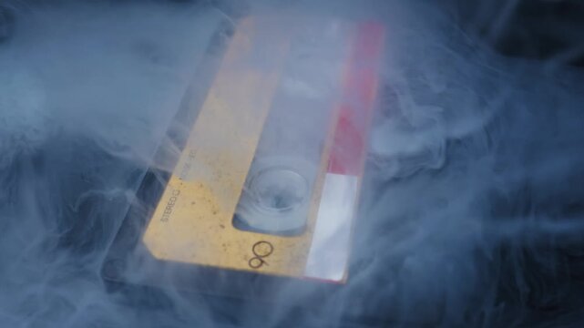 Vintage audio cassette tape with a weathered yellow label is engulfed by white smoke. Close-up shot on a dark background with atmospheric, moody lighting.