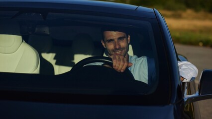 Handsome successful entrepreneur looking through windshield of modern electric car. Portrait of young smiling businessman in his innovative electrical vehicle. Slow motion