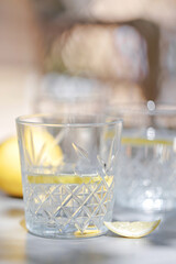 Detailed close-up of a textured crystal glass with lemon water on a gray table, surrounded by citrus fruit and dappled light