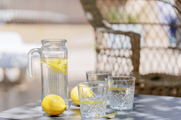 Wide shot of a summer outdoor table with glass pitcher of lemon water, three glasses, and whole lemons with blurred natural background