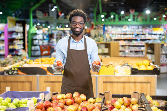 Smiling male grocer wearing an apron and gloves, standing in a supermarket produce section, holding two red apples, and looking at the camera with a friendly expression