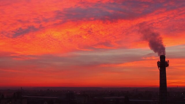 Industrial chimney emitting smoke against a fiery sunrise sky. Air pollution and environmental concept during winter dawn. orange sunrise sky