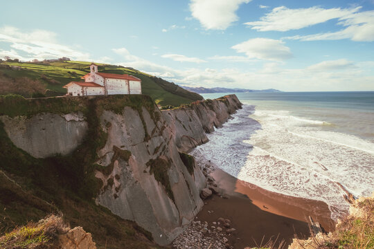 View of a church perched atop jagged cliffs where the land meets the turbulent sea under a vast sky, creating a serene yet dramatic coastal panorama, Zumaia, Basque Country, Spain.