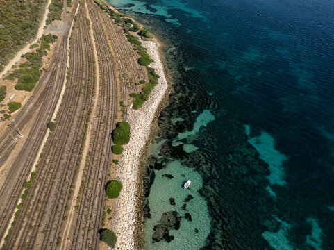 Aerial view of turquoise waters meeting the rugged coastline, contrasted against the parallel lines of the railway tracks cutting through the arid landscape, Sardinia, Italy.