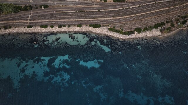 Aerial view of the coastline where the turquoise sea meets the rough rocky shore, with a railway line running parallel, Cagliari, Sardinia, Italy.