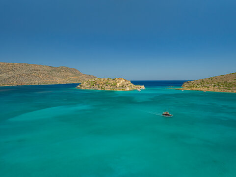 Aerial view of a small boat sailing on the turquoise waters towards the island of Spinalonga with its historic ruins, Spinalonga, Lasithi, Greece.
