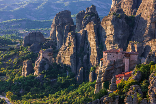 Aerial view of monasteries perched atop towering rock formations, bathed in golden light and surrounded by lush greenery, Kalabak, Trikala, Greece.