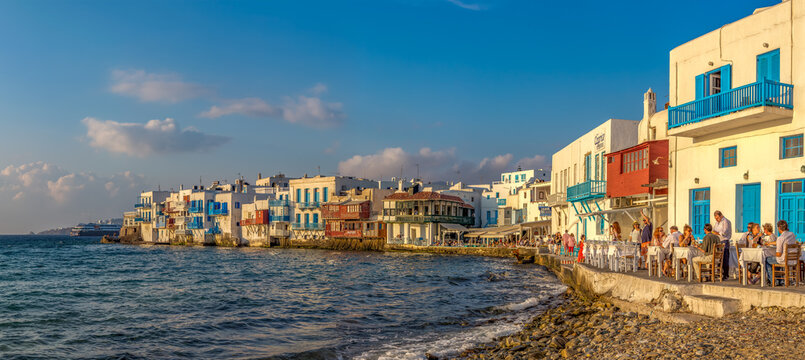 Aerial view of whitewashed buildings with vibrant blue accents hug the coastline, as people dine al fresco against the backdrop of the Aegean Sea, Mykonos, Greece.