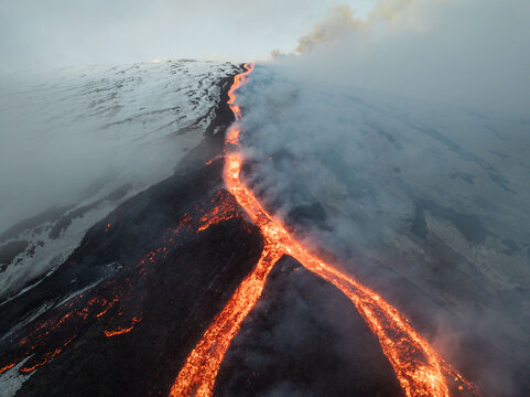 Aerial view of fiery lava cascading down the snow-dusted slopes of Mount Etna, a dramatic display of nature's power amidst the clouds, Catania, Sicily, Italy.