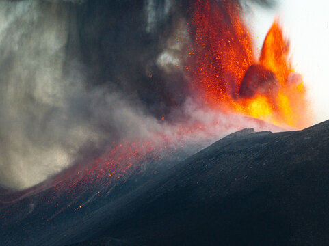Aerial view of fiery lava cascading down the dark slopes of Mount Etna, a volcanic spectacle of intense heat and dramatic smoky plumes, Catania, Sicily, Italy.
