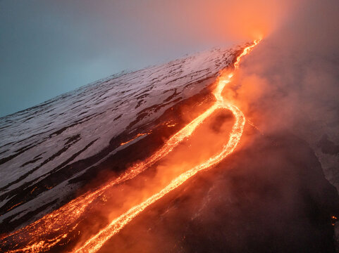 Aerial view of fiery lava flowing down the snow-covered slopes of Mount Etna, a stark contrast between the molten orange and the icy white, Catania, Sicily, Italy.