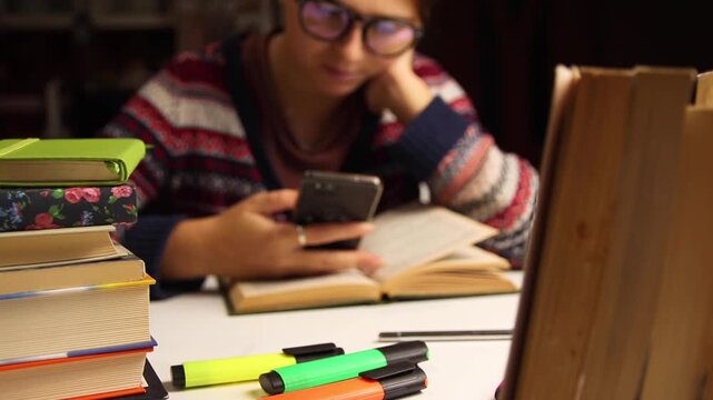 Young woman student in glasses sitting at a desk with a stack of books and using a smartphone. Procrastination, online learning, and education concept
