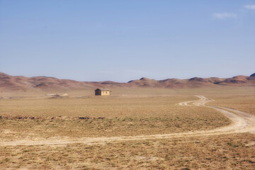 Obraz premium A lonely abandoned village in the endless steppe of Kazakhstan. A desert landscape with a winding dirt road leading to an old building set against a backdrop of low hills. 