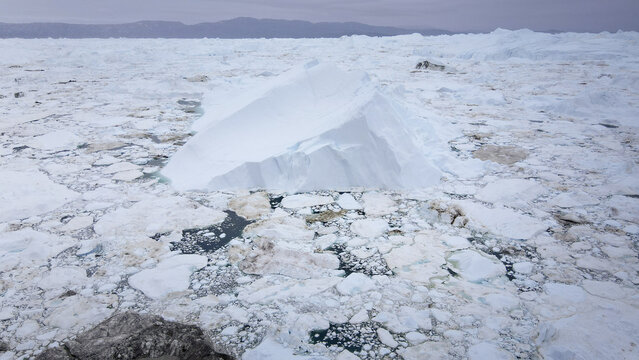 Aerial view of a pristine white iceberg rises majestically from the fragmented ice floes, creating a stunning contrast against the distant, muted coastline, Greenland, Greenland.