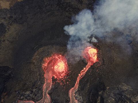 Aerial view of the fiery, molten lava flowing down the dark, rugged terrain, contrasting with the billowing white smoke, Grindavik, Grindavikurbaer, Iceland.