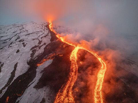 Aerial view of a fiery river of lava snaking down the snow-dusted slopes of Mount Etna, a stark contrast against the cool, misty sky, Catania, Sicily, Italy.