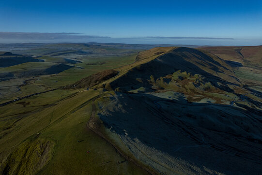 Aerial view of sun-kissed Mam Tor ridge casting long shadows over verdant fields, a tapestry of green and gold under a crisp blue sky, Hope Valley, England, United Kingdom.