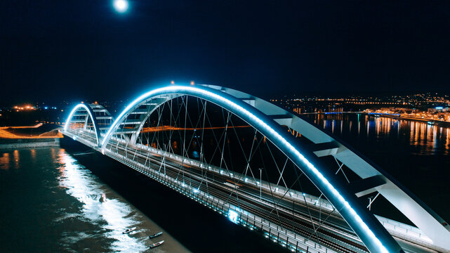 Aerial view of illuminated bridge arches mirroring in the dark water, a modern marvel under the moon's glow, Novi Sad, Vojvodina, Serbia.