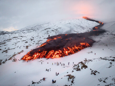 Aerial view of a fiery river of molten lava snaking down the snow-covered slopes of Mount Etna, a stark contrast of intense heat and icy cold, Catania, Sicily, Italy.