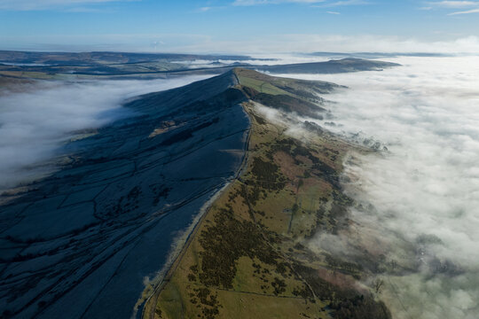 Aerial view of Mam Tor's ridge cuts through a sea of mist, the sunlit slopes contrasting with the shadowed valleys, Hope Valley, England, United Kingdom.