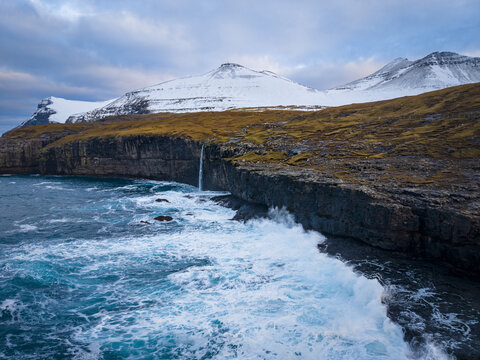 Aerial view of a waterfall cascading from rugged cliffs into the turbulent ocean, with snow-capped mountains under a cloudy sky, Eidi, Eysturoy, Faroe Islands.