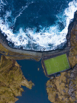 Aerial view of a solitary football field, a vibrant green rectangle nestled between the rugged coastline and the turbulent North Atlantic, Eidi, Eysturoy, Faroe Islands.