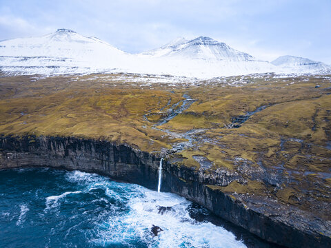 Aerial view of a dramatic waterfall cascading into the turbulent ocean, framed by rugged cliffs and snow-dusted peaks, Eidi, Eysturoy, Faroe Islands.