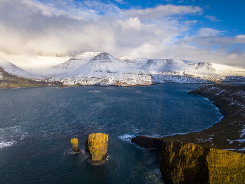 Aerial view of the dramatic Drangarnir sea stacks piercing the turquoise waters against the backdrop of snow-dusted mountains, Drangarnir, Faroe Islands.