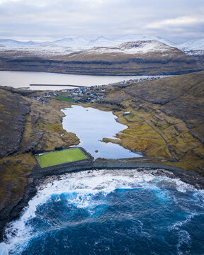 Aerial view of where rugged coastline meets the tranquil lake near a vibrant green football field, Eidi, Eysturoy, Faroe Islands.
