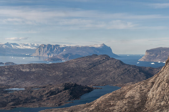 Aerial view of rugged, rocky terrain meets the frigid sea, dotted with icebergs, against a backdrop of towering, snow-capped mountains, Greenland.