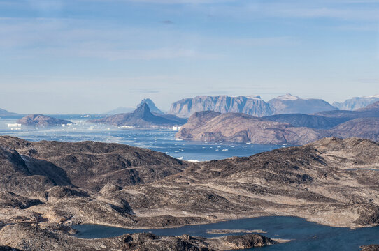 Aerial view of rugged, rocky terrain meets the icy expanse of the sea, punctuated by scattered icebergs and distant, majestic mountains, Nuuk, Greenland.