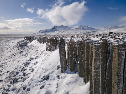 Aerial view of striking basalt columns stand sentinel against a backdrop of snow-dusted mountains under a sky brushed with wispy clouds, Raudhamelur, Eyja- og Miklaholtshreppur, Iceland.