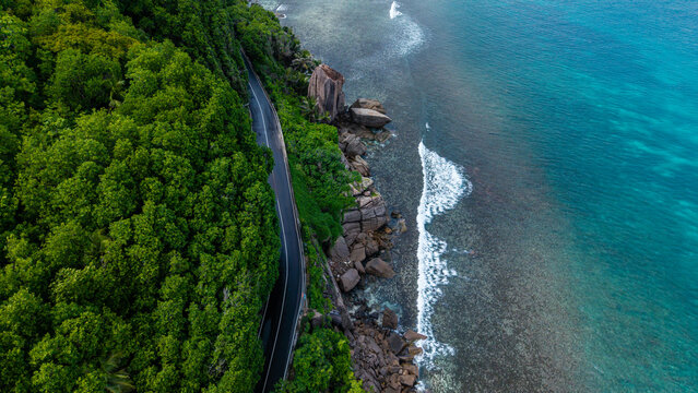 Aerial view of the dark road winding along the vibrant green forest edge and the turquoise ocean near Anse Forbans, Mahe, Seychelles.