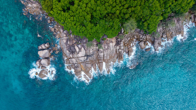 Aerial view of the dramatic coastline where verdant jungle meets the textured rocks, kissed by foamy waves in the turquoise sea, Takamaka, Seychelles.