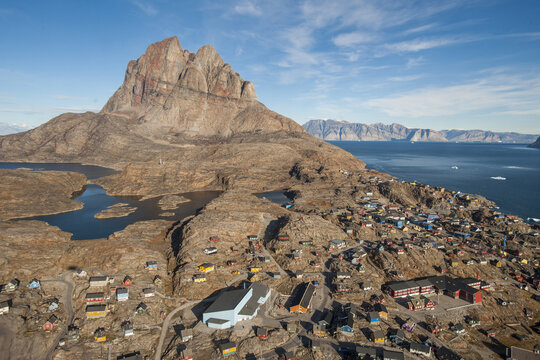 Aerial view of rugged mountains meet colorful dwellings nestled on the rocky coast, where ice-dotted waters stretch to distant peaks, Tasiilaq, Greenland.