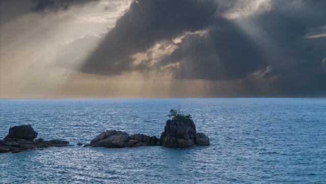 Aerial view of sunbeams piercing through dark clouds, illuminating the tranquil turquoise waters around the rocky outcrop of Anse Soleil, Mahe, Seychelles.