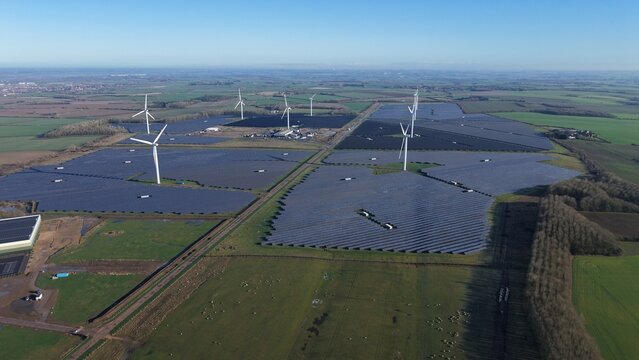 Aerial view of wind turbines standing tall amidst rows of solar panels, creating a stark contrast against the green fields, Chelverston, Wellingborough, United Kingdom.